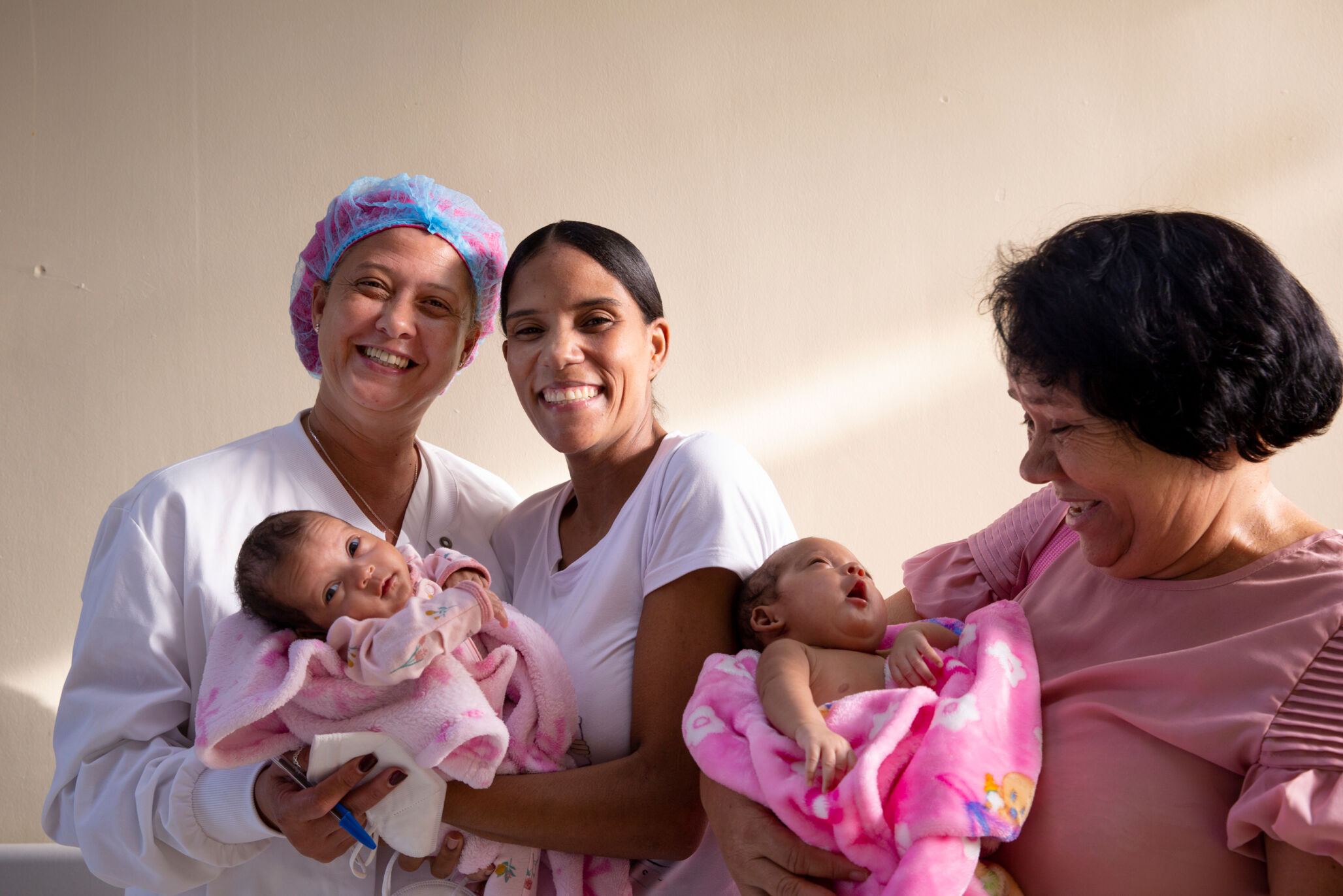 A new mother smiles while holding her baby, along with medical staff. Another woman holds a newborn and smiles.
